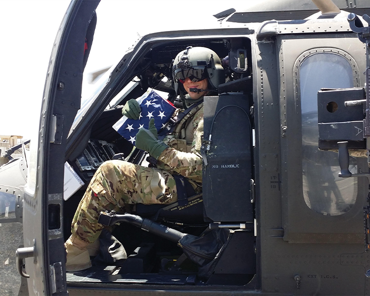 A SOLIDER IN A MILITARY HELICOPTER HOLDS A FOLDED FLAG AND GIVES A THUMBS-UP SIGN